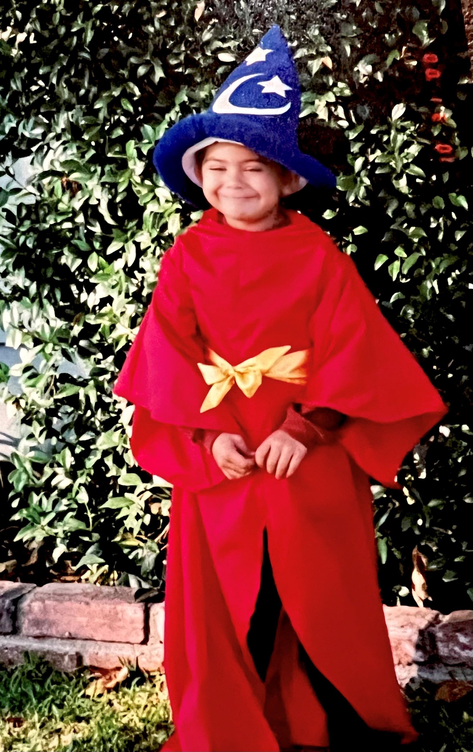 A young boy smiles at the camera. He is dressed in a red rob and wizard hat.