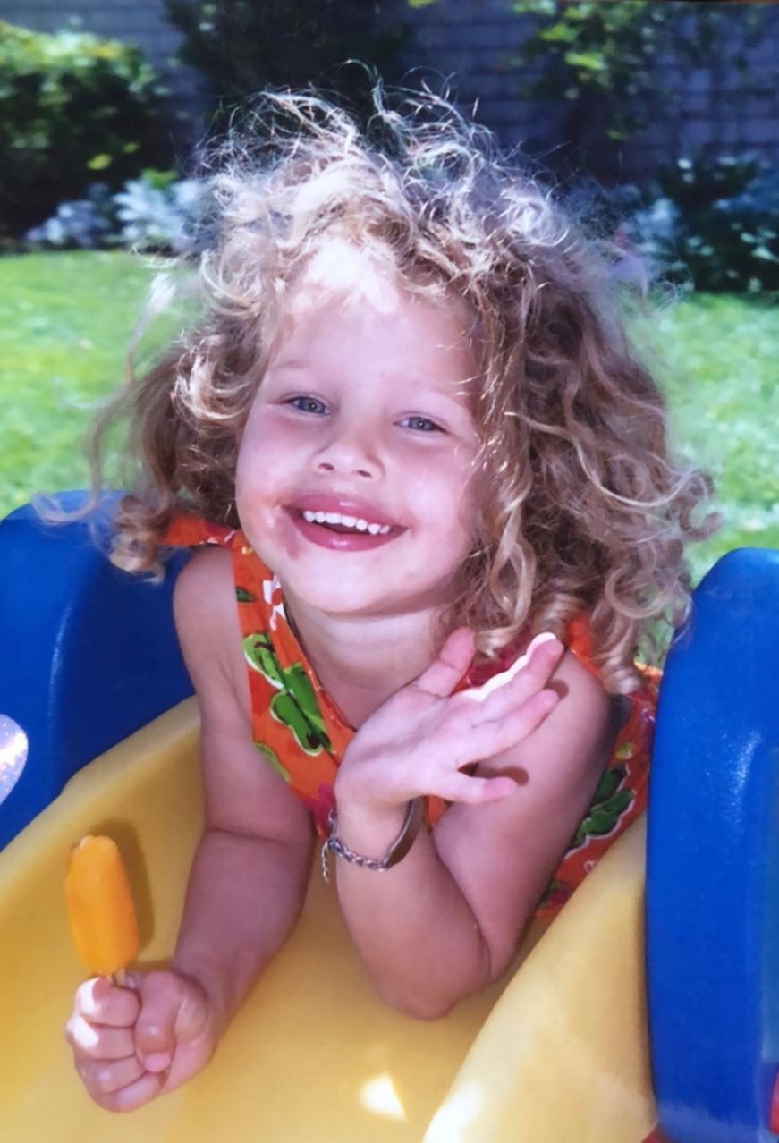 A young girl with curly blond hair smiles at the camera. She's eating a popsicle.
